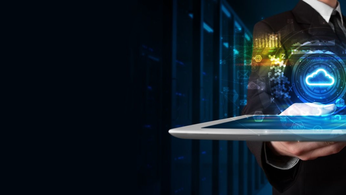 A person in a suit holding a tablet with a glowing cloud computing icon and digital data graphics hovering above it, symbolizing cloud technology and data management, with a dark server room background.