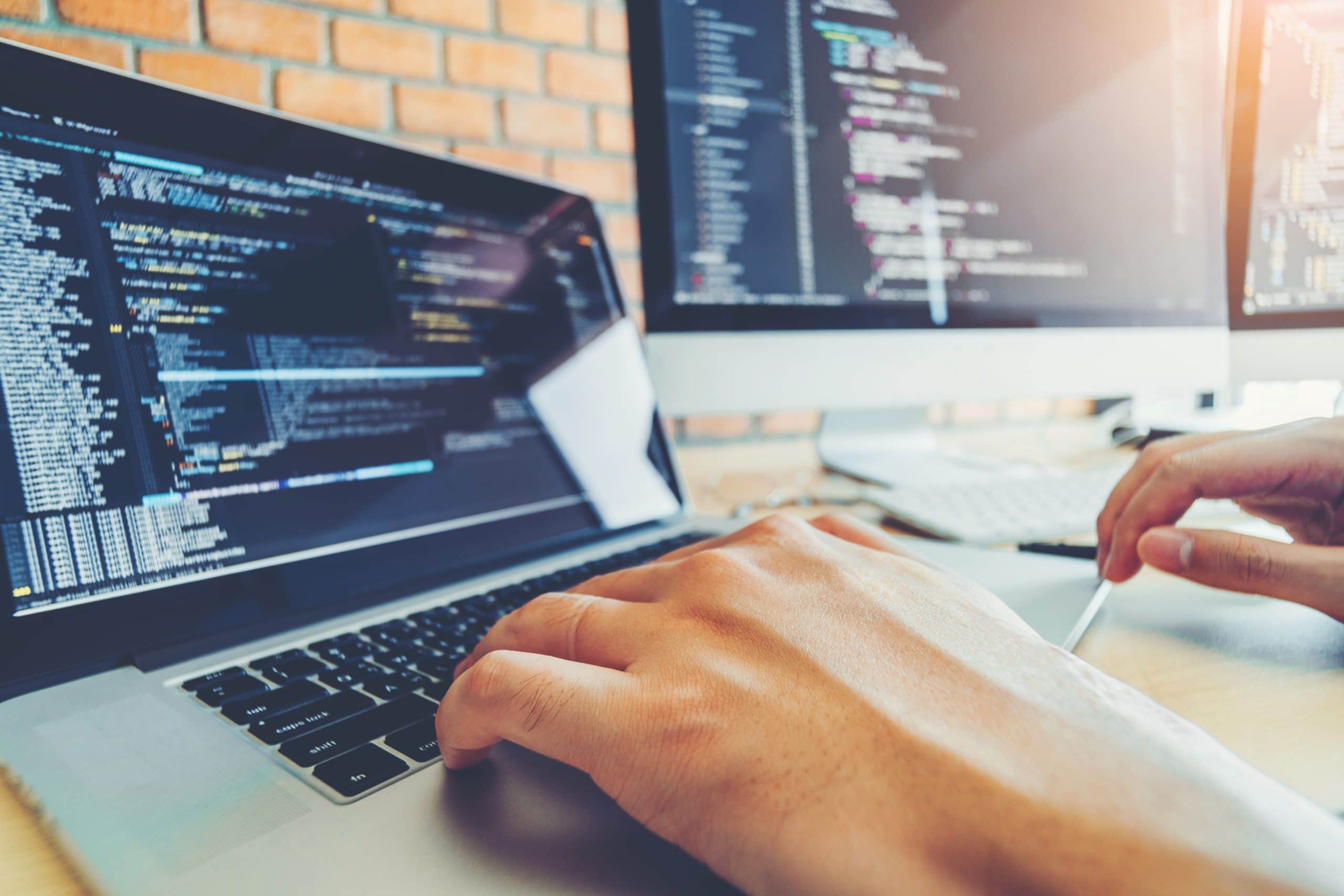 A person types on a laptop with programming code visible on the screen, possibly working on enVista Careers or a parcel audit service. Another monitor in the bright workspace, set against a brick wall, also displays code.