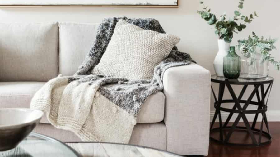 A light beige sofa with a chunky knit throw blanket and pillow sits next to a round black side table holding green glass vases and leafy plants, creating a cozy, modern living room setting.