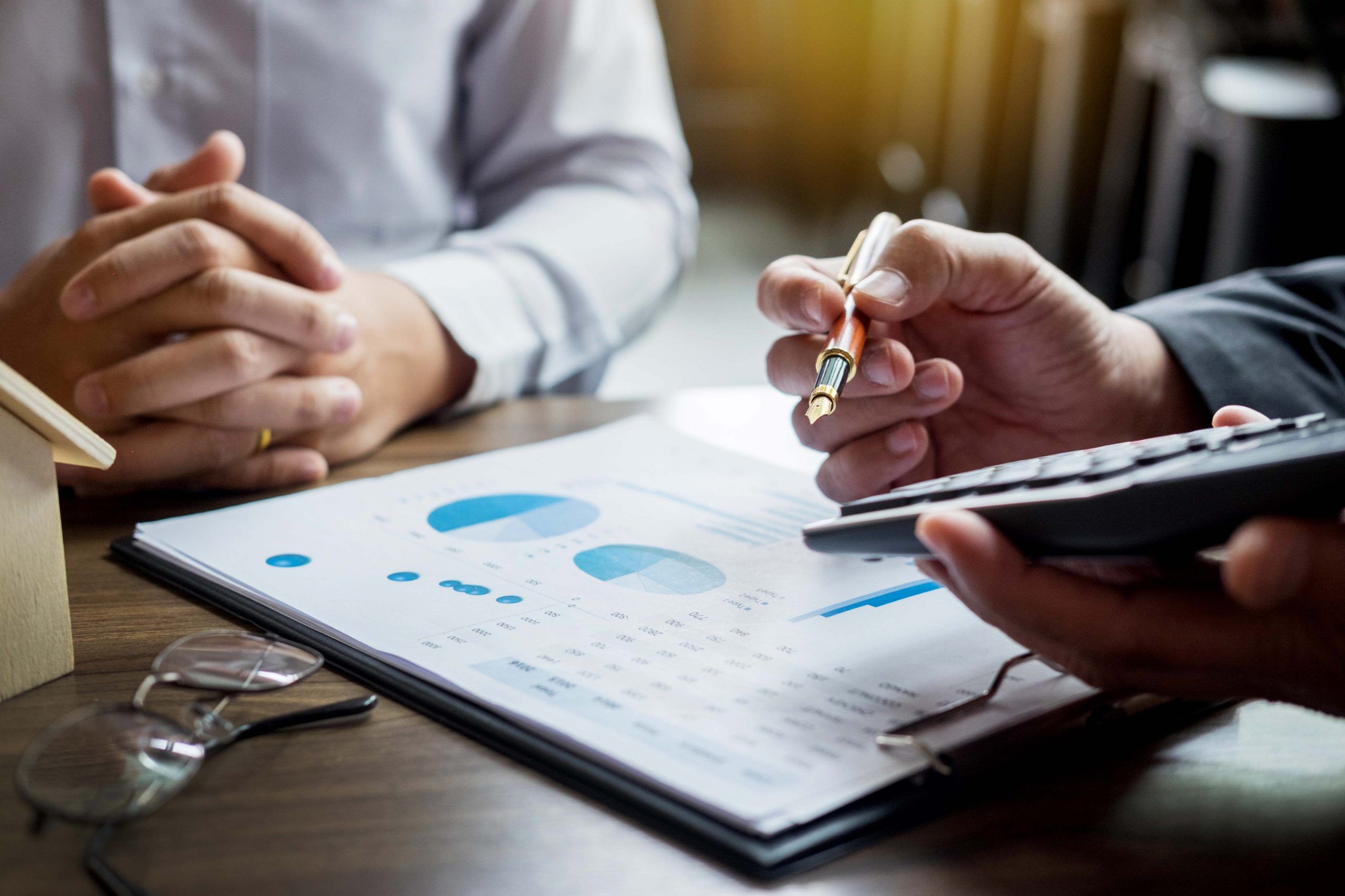 Two people sit at a desk reviewing financial charts on a clipboard, possibly discussing enVista 365 pricing. One holds a pen and calculator, while the other rests their hands together. Glasses and a small house model are also on the table.