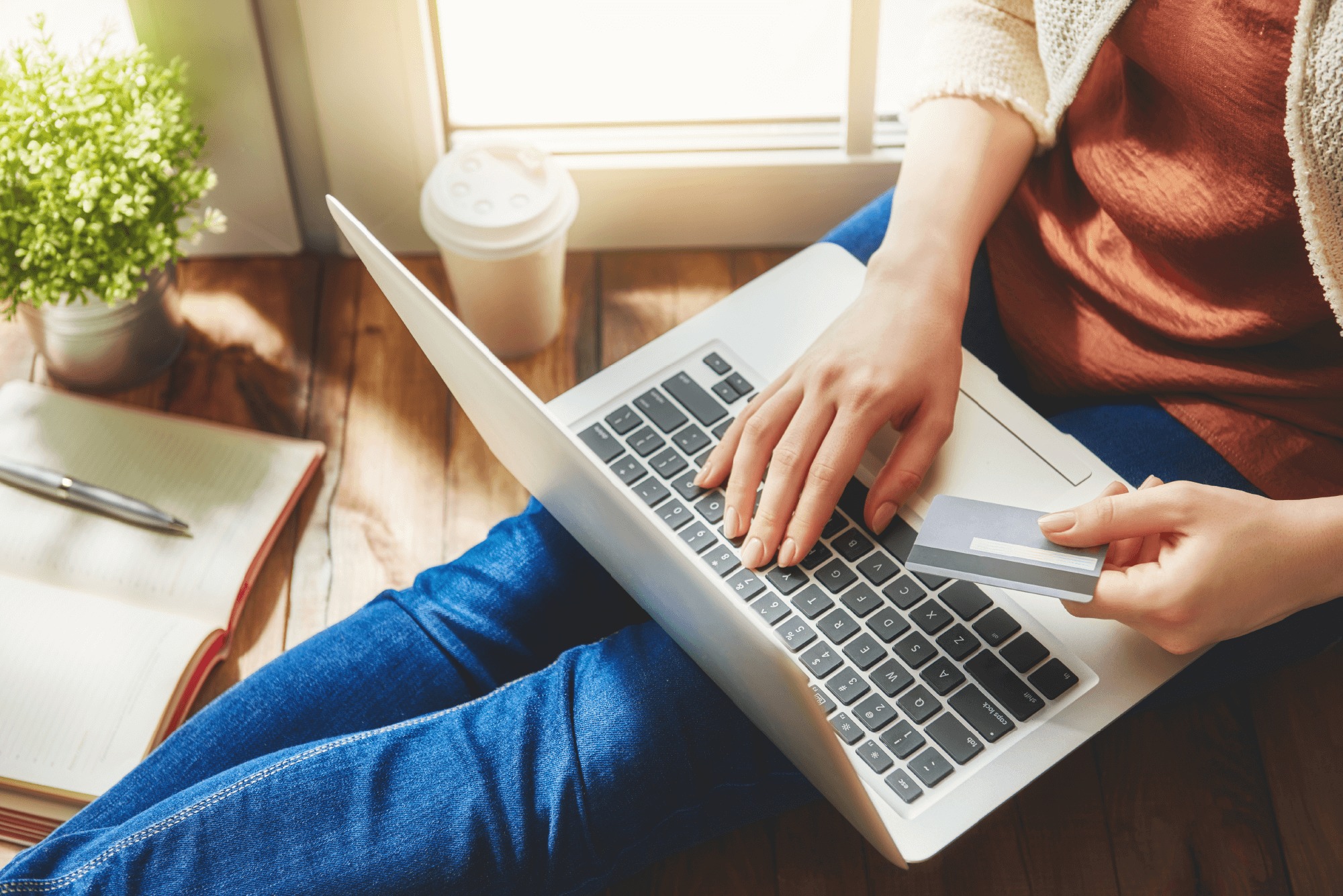 A person sitting on the floor with a laptop on their lap, holding a credit card, shopping online. A notebook, coffee cup, and potted plant are nearby. Sunlight shines through a window.