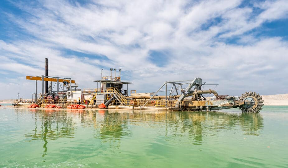 A large industrial dredging machine floats on green water under a partly cloudy blue sky, with pipes and equipment visible on its deck.