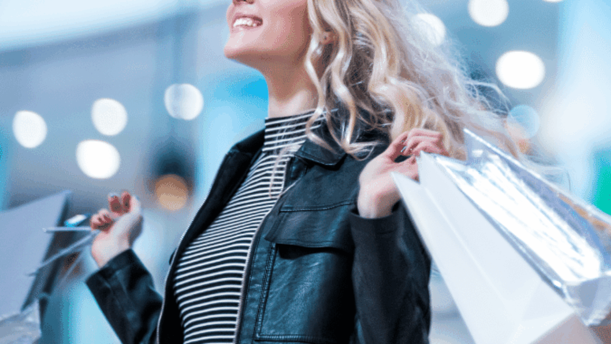 A smiling woman with long blonde hair, wearing a black leather jacket and striped shirt, holds several shopping bags in both hands. The background is blurred with bright lights, suggesting a mall or shopping center.