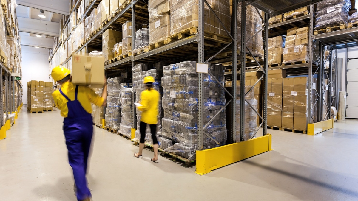 Two workers in safety helmets and uniforms move boxes in a large warehouse with tall shelves stacked with boxes and pallets. The warehouse is brightly lit and organized, with wide aisles for easy movement.