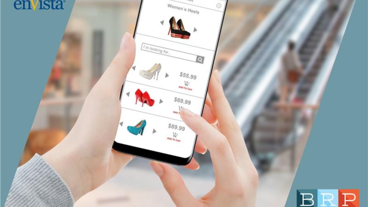 A person holds a smartphone displaying an online shopping app for women’s heels, with several shoe options and prices. An escalator and blurred shopping mall background are visible.