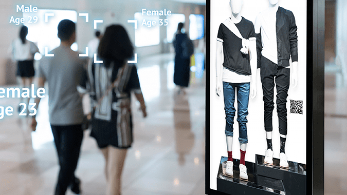 A digital billboard in a mall displays mannequins in casual clothing. Nearby, a group of people walks past, while facial recognition technology identifies their gender and estimated age on screen overlays.
