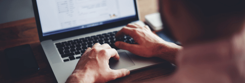 A person is typing on a laptop keyboard at a wooden desk. The screen displays a document or webpage, and a smartphone lies nearby. The scene suggests working or studying.