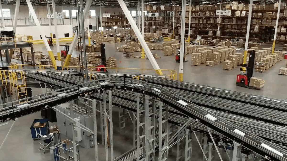 Large warehouse interior with rows of shelves stacked with boxes, several forklifts moving pallets, and a network of automated conveyor belts in the foreground. The space is well-lit and organized for distribution.