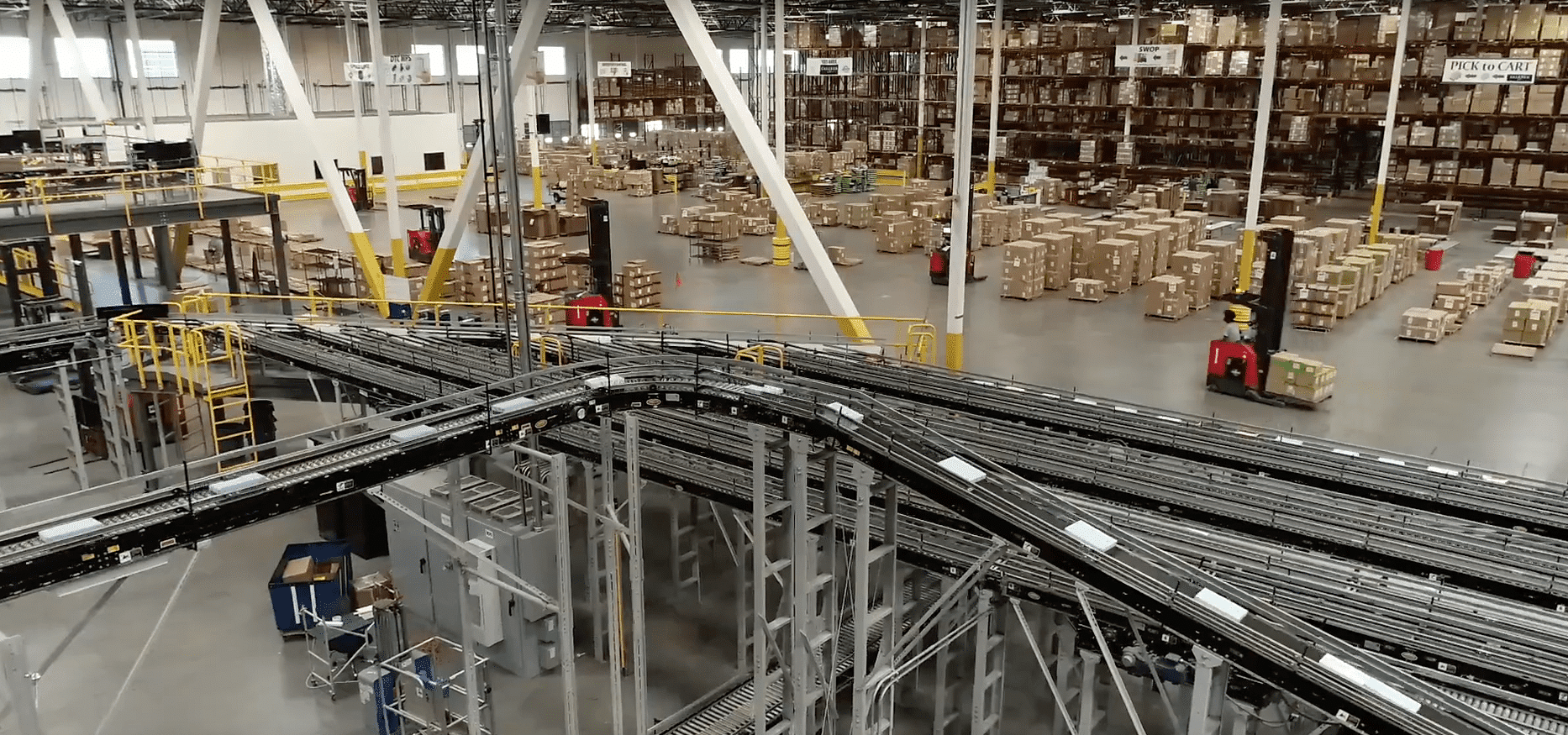 Large warehouse interior with rows of shelves stacked with boxes, several forklifts moving pallets, and a network of automated conveyor belts in the foreground. The space is well-lit and organized for distribution.