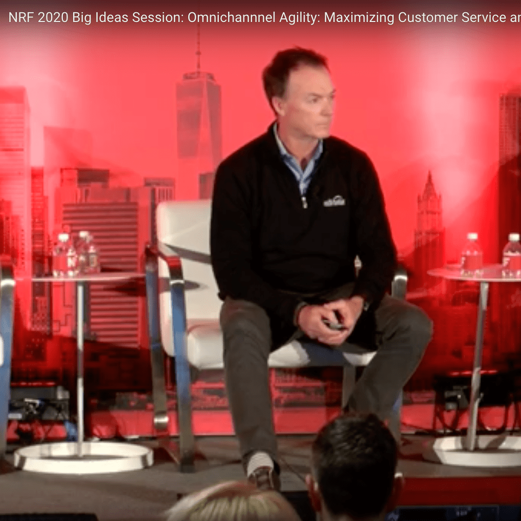 A man in a dark jacket and gray pants sits on stage in front of a red backdrop with cityscape graphics during the NRF 2020 Big Ideas Session. He is holding a microphone and appears to be listening.