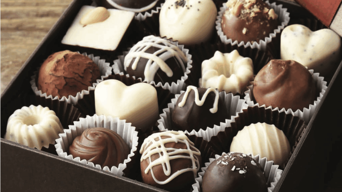 An open box of assorted chocolates, including heart-shaped, round, and square pieces in dark, milk, and white chocolate, displayed neatly in paper cups on a wooden surface.