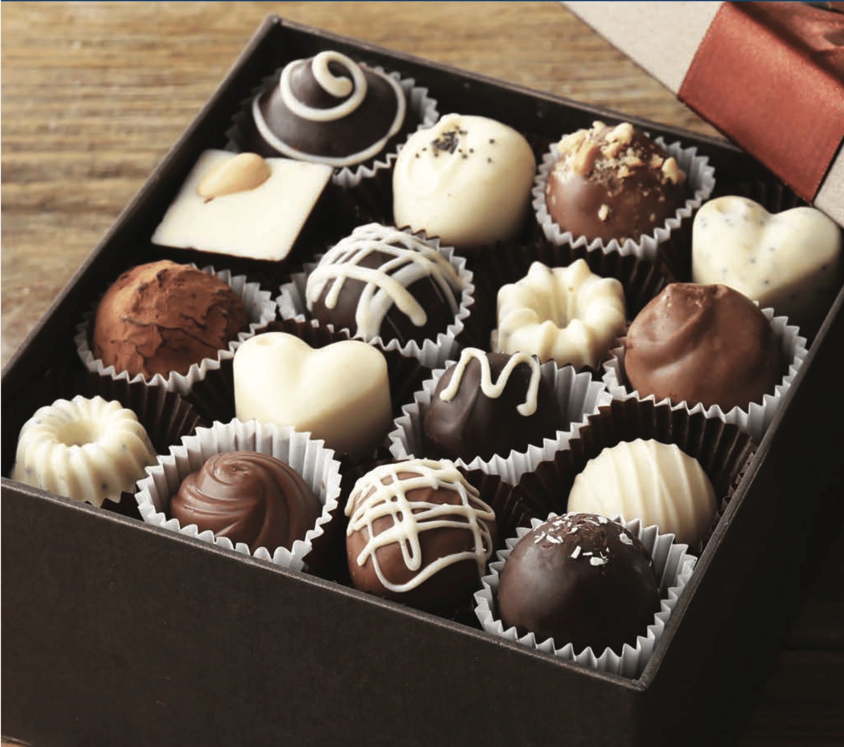 An open box of assorted chocolates, including heart-shaped, round, and square pieces in dark, milk, and white chocolate, displayed neatly in paper cups on a wooden surface.