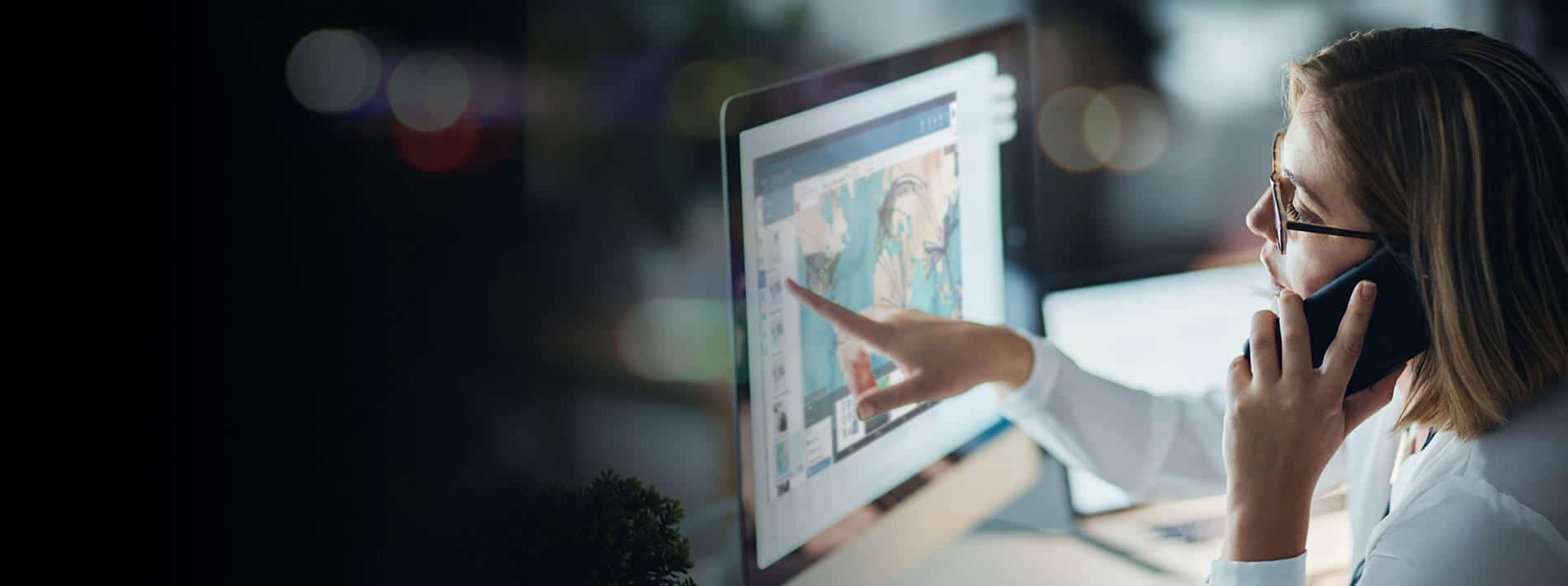 A woman wearing glasses talks on the phone while pointing at a computer screen displaying colorful graphics or maps in a dimly lit office environment.