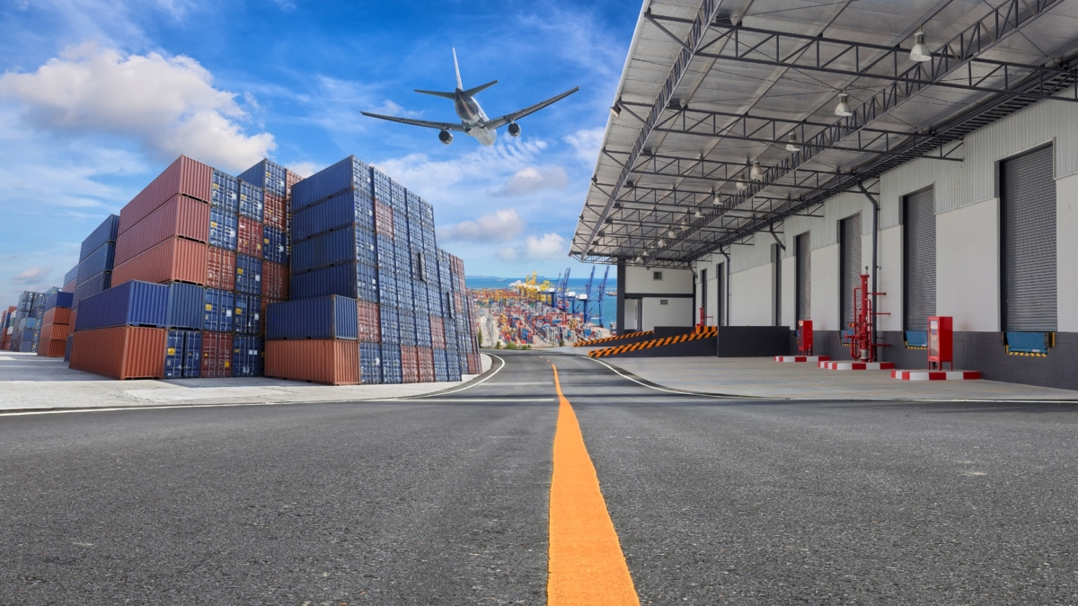 A cargo plane flies over stacked shipping containers near a warehouse at an industrial port, with a clear sky and road marking leading toward the buildings.