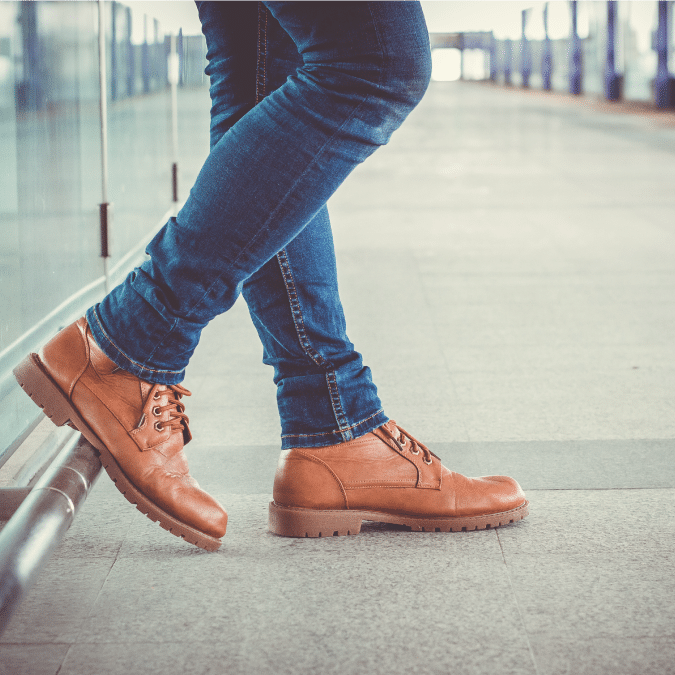 Person wearing blue jeans and tan leather boots, standing with crossed legs on a smooth indoor floor near a glass wall, with a blurred background suggesting a spacious, modern building or hallway.