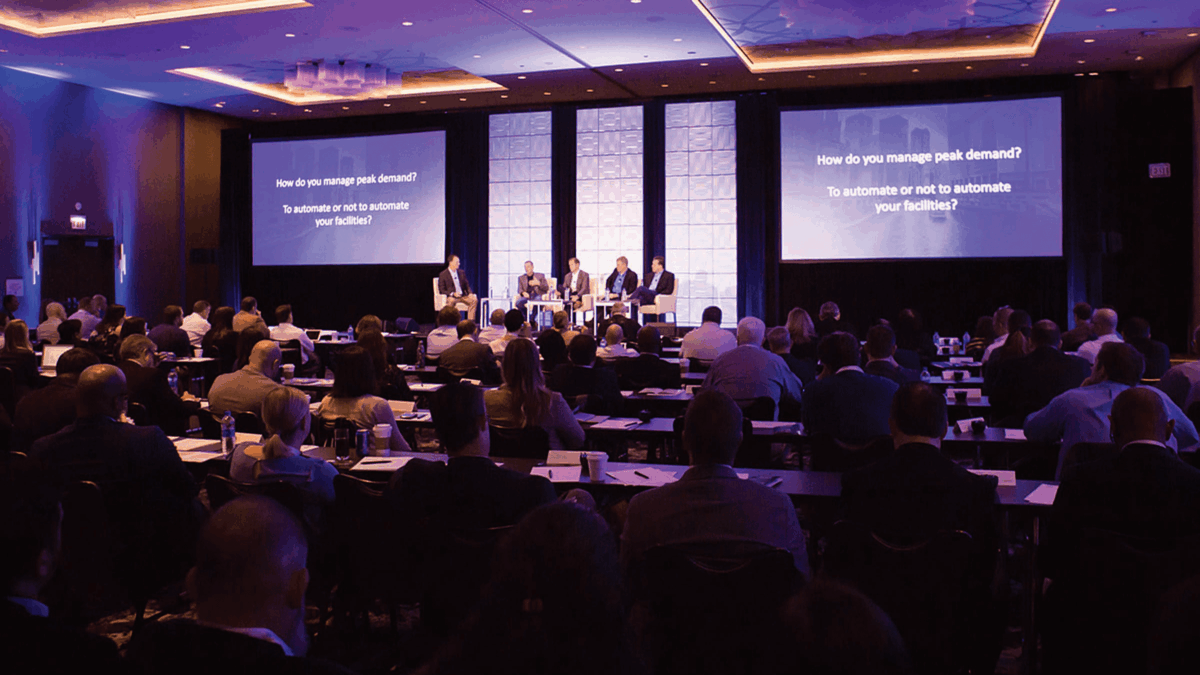 A large audience watches a panel discussion in a conference room. Four speakers sit onstage in front of three large screens displaying the question, “How do you manage peak demand? To automate or not to automate your facilities?”.
