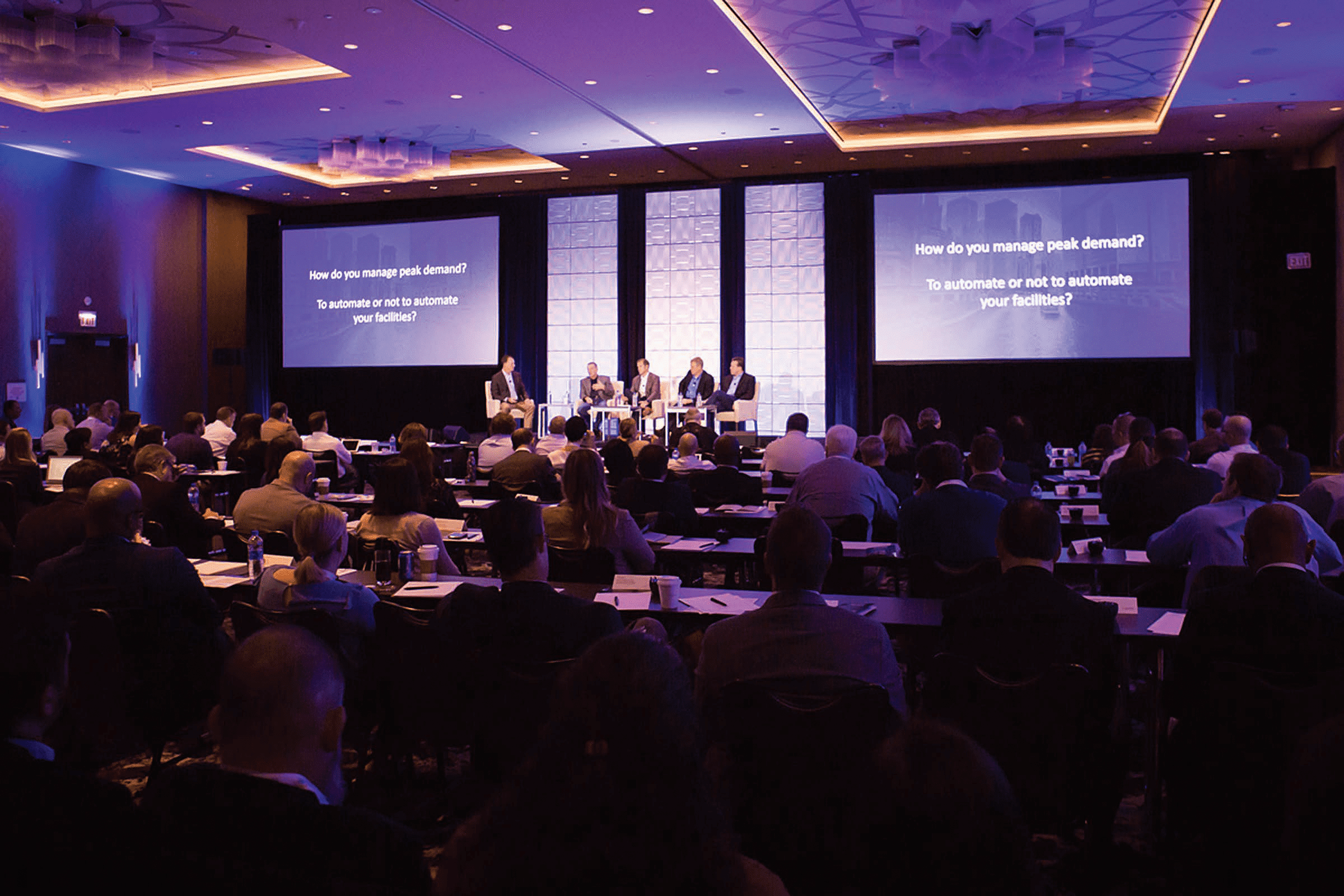 A large audience watches a panel discussion in a conference room. Four speakers sit onstage in front of three large screens displaying the question, “How do you manage peak demand? To automate or not to automate your facilities?”.