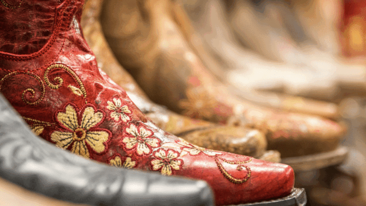 Close-up of an ornate red cowboy boot with yellow floral embroidery, displayed among several other brown and tan cowboy boots in the background on a shelf.