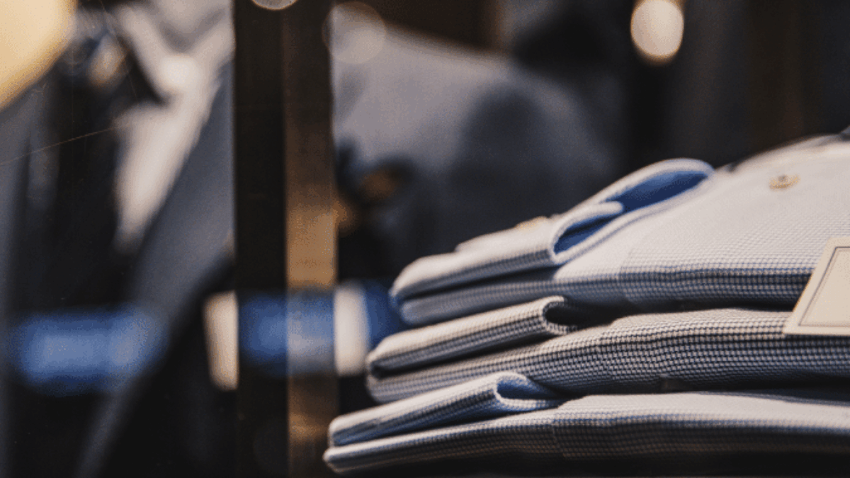 A close-up of neatly folded blue dress shirts displayed on a shelf in a clothing store, with blurred suits and garments in the background.