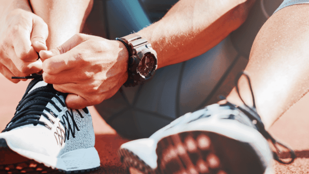 Close-up of a person in athletic clothing sitting on a red running track, tying the laces of their white and black running shoes. Sunlight highlights their arm and wristwatch.