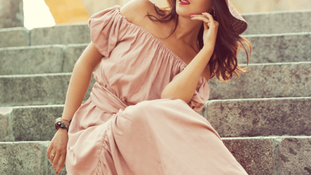 A woman in a pink off-shoulder dress and wide-brimmed hat sits gracefully on stone steps, gazing to the side with a thoughtful expression. She wears sandals and has her hair loose, with a background of stone architecture.