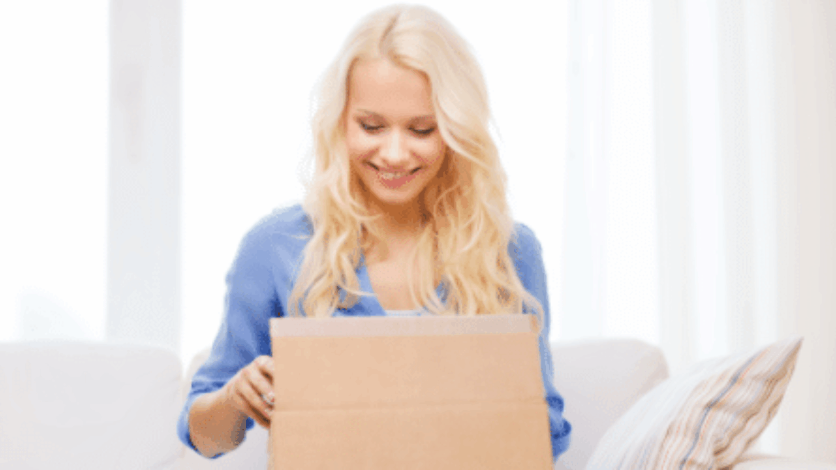A smiling woman with long blonde hair sits on a couch and opens a cardboard box, looking inside with excitement. A striped pillow is beside her, and soft daylight fills the room.