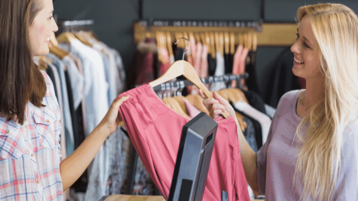 Two women stand at a clothing store counter, smiling as one hands the other a pink garment on a hanger, with shelves of folded clothes and hanging outfits in the background.