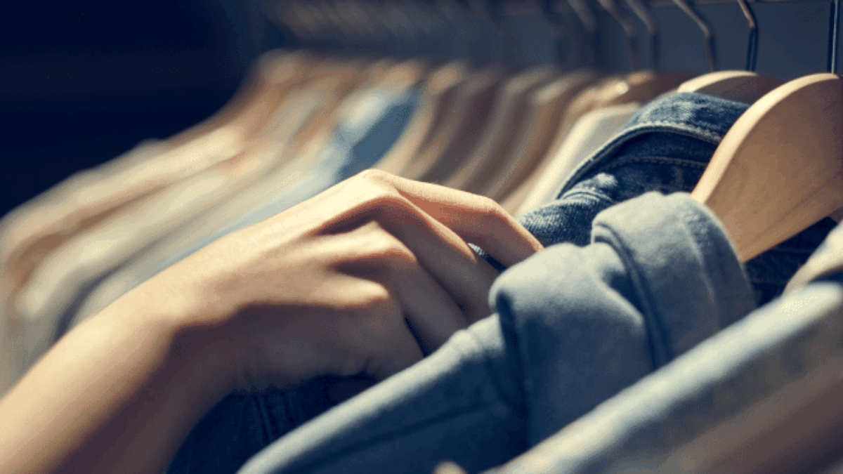 A close-up of a person’s hand browsing through clothes on hangers in a store, with soft lighting highlighting the fabrics and different shades of denim and cotton shirts.