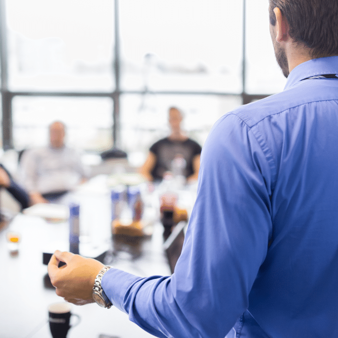 A person in a blue shirt stands and gestures while giving a presentation to a group of people seated around a conference table in a bright, modern office setting.