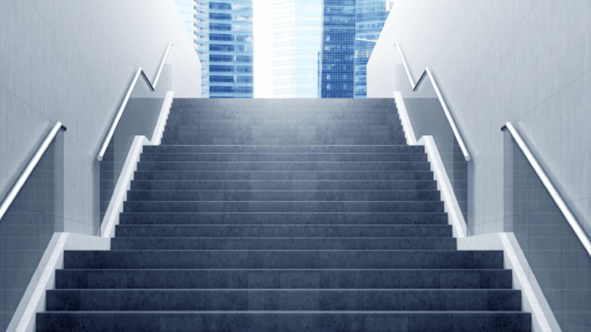 A wide, modern staircase with metal railings on both sides leads upward to bright daylight, with tall glass office buildings visible in the background.