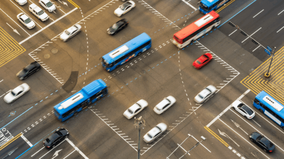Aerial view of a busy city intersection with cars, buses, and a red vehicle moving in multiple directions, traffic lanes and markings clearly visible.