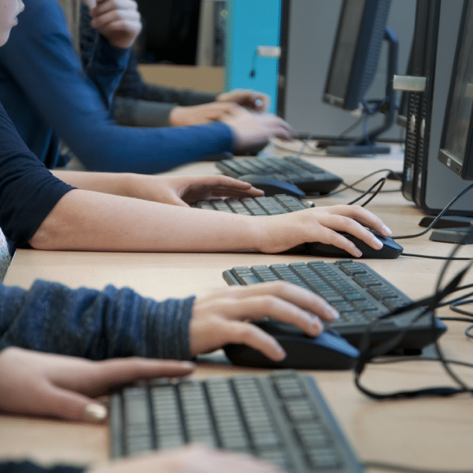 Several people sit at a row of computers, typing on keyboards and using mice. Only their hands and arms are visible, suggesting a classroom or computer lab setting.