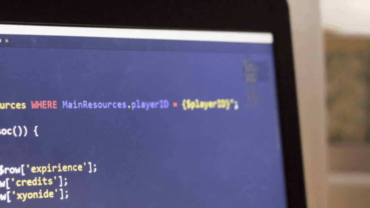 A close-up of a computer screen displaying code, with part of a laptop bezel visible. The code appears to be written in SQL and another programming language. Blurred curtains and sunlight are in the background.