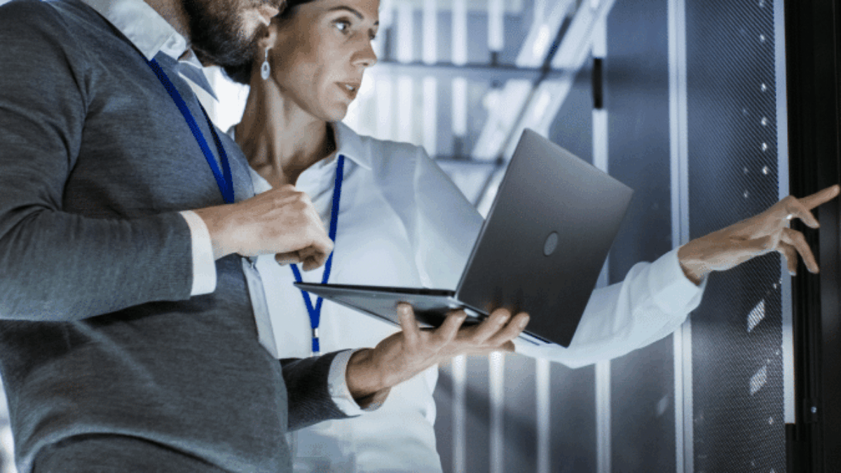 Two IT professionals in a server room, one holding a laptop and the other pointing at a server rack, both wearing ID badges and focused on their work.