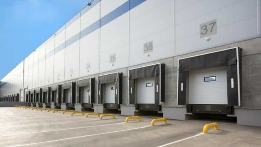 A row of loading docks with numbered bays and closed overhead doors at a large, modern warehouse under a clear blue sky.