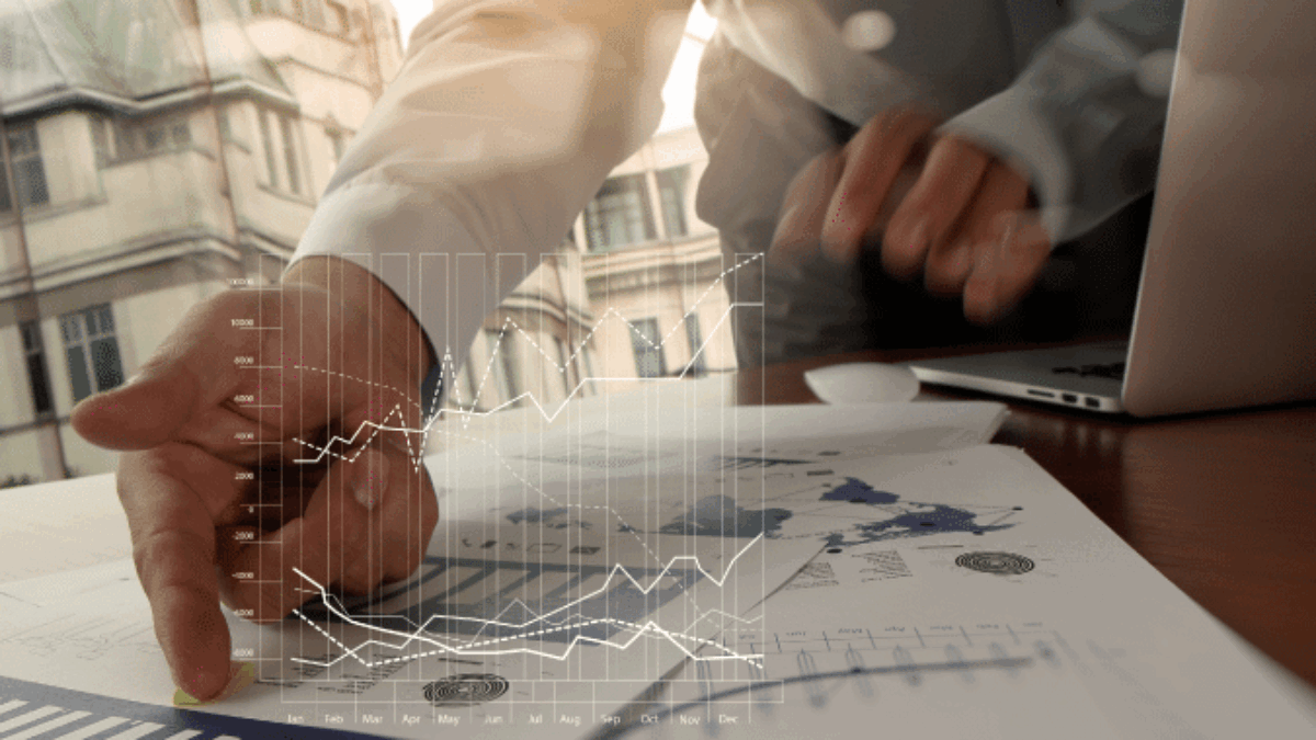 A person points at financial documents and graphs on a desk, with a laptop nearby and a virtual graph overlay, suggesting data analysis or business strategy in a modern office setting.