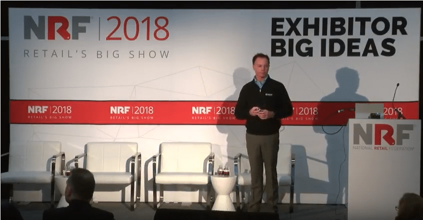 A man stands on stage giving a presentation at the NRF 2018 Retail’s Big Show, with “Exhibitor Big Ideas” displayed behind him and empty white chairs beside him.