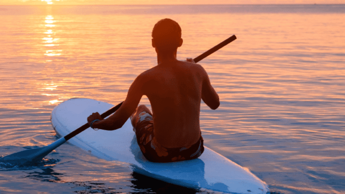 A person paddles on a stand-up paddleboard in calm ocean waters at sunset, with warm orange and pink hues in the sky and sunlight reflecting on the water’s surface.