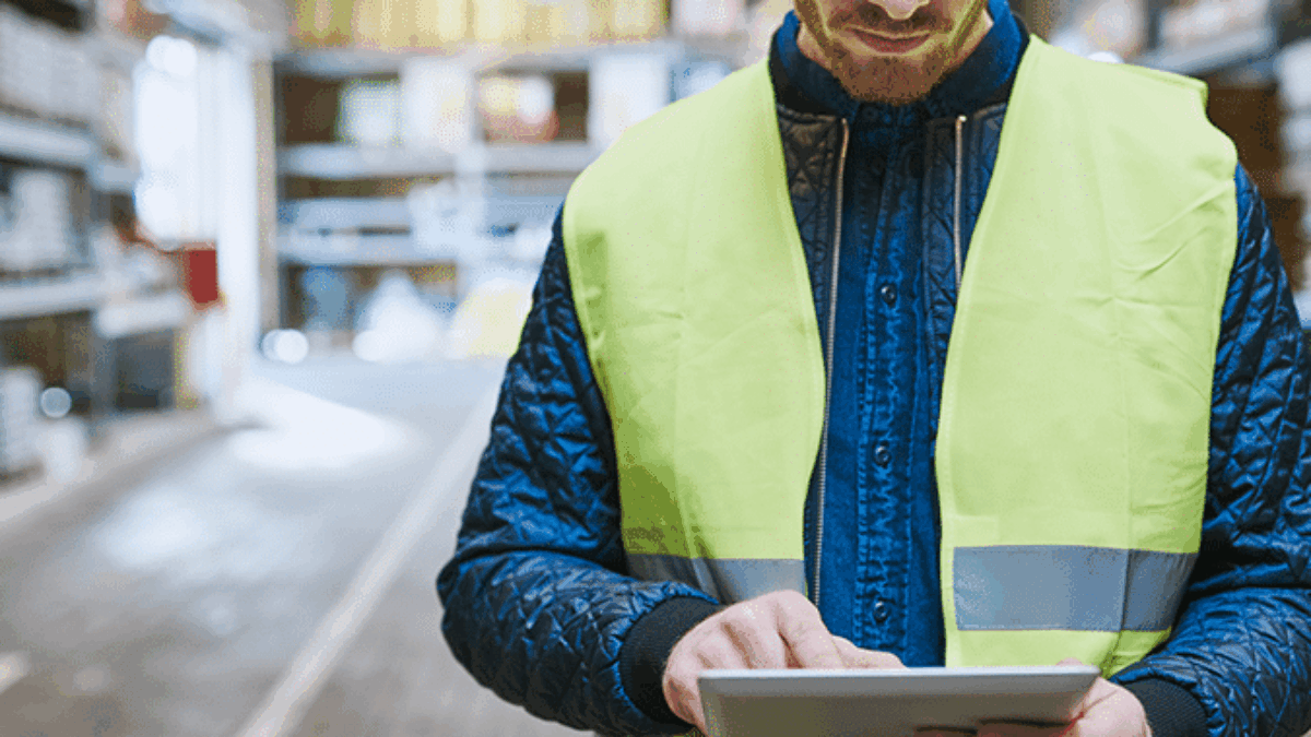 A man wearing a yellow safety vest stands in a warehouse, looking down and using a tablet. Shelves with goods are visible in the background, and the setting appears organized and well-lit.