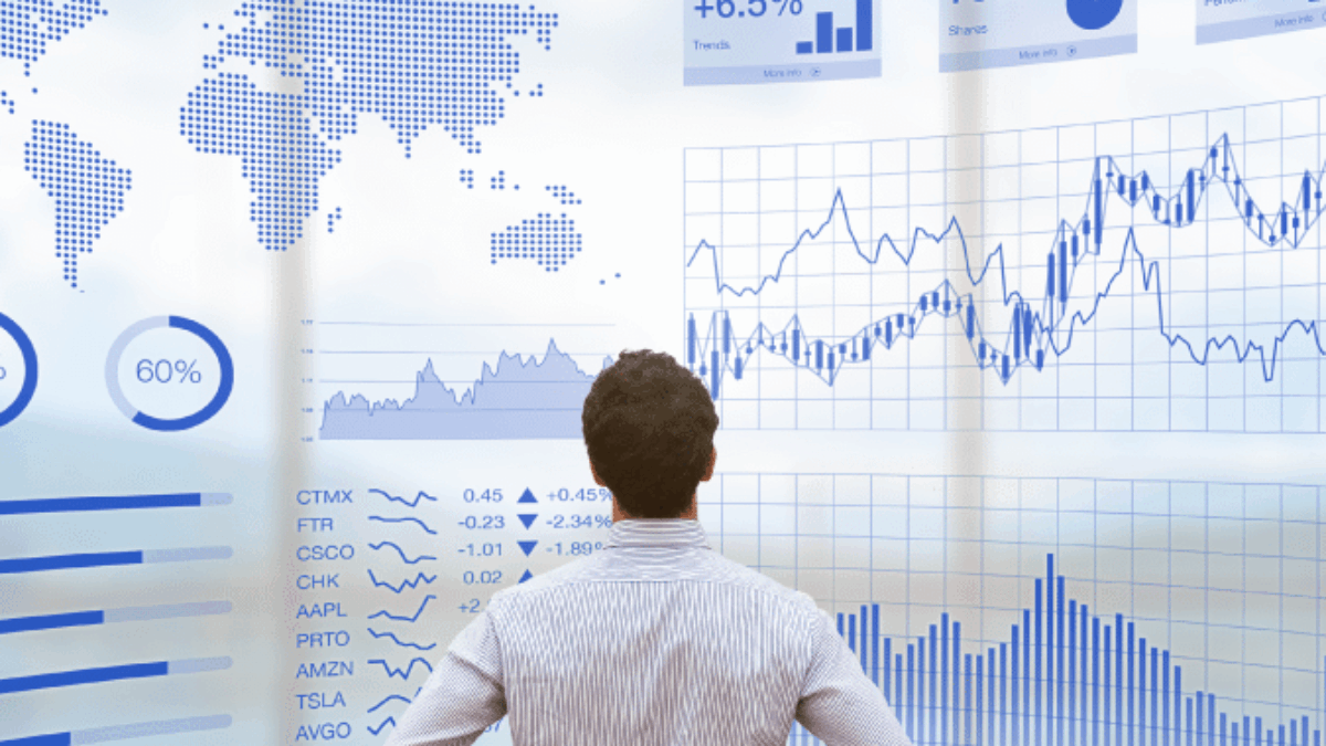 A man stands with hands on hips, facing a large digital screen displaying world maps, stock market data, charts, graphs, and various financial statistics in blue tones.