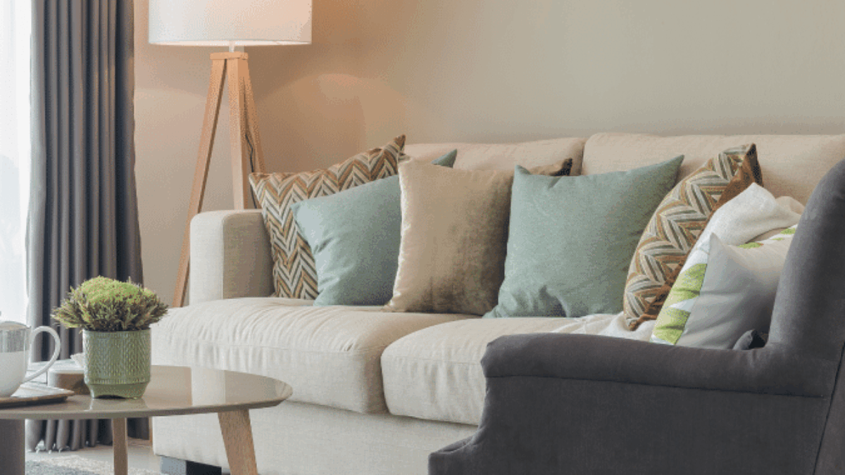 A cozy living room with a beige sofa decorated with green and patterned pillows, a dark gray armchair, a wooden coffee table with a small plant and mugs, a standing lamp, and floor-length curtains.