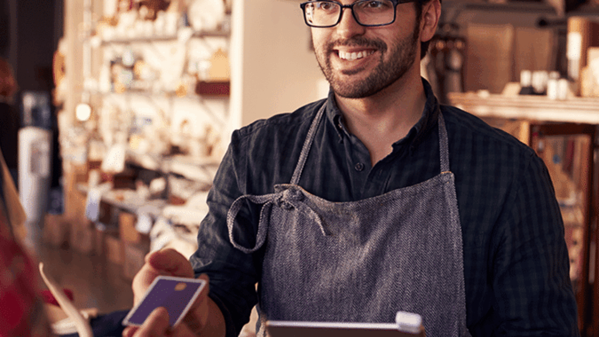 A smiling shop employee wearing an apron and glasses holds a tablet while a customer hands him a credit card in a cozy, well-lit store.