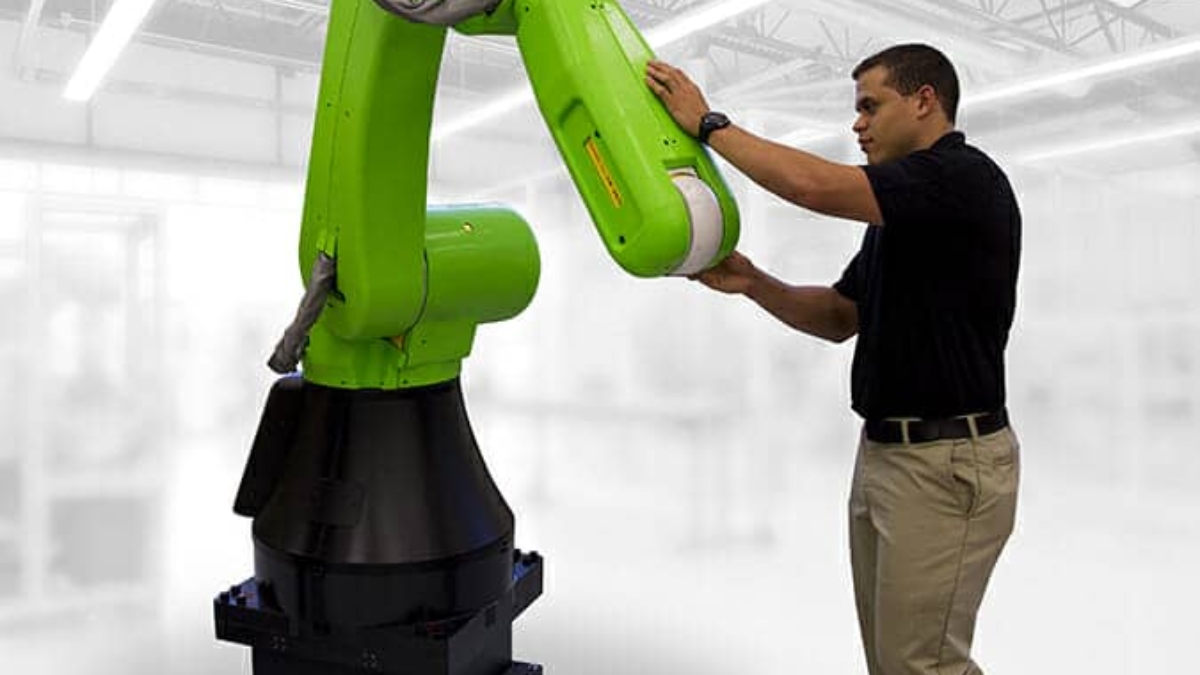 A man in khaki pants and a black shirt interacts with a large green industrial robotic arm in a bright, modern factory setting.