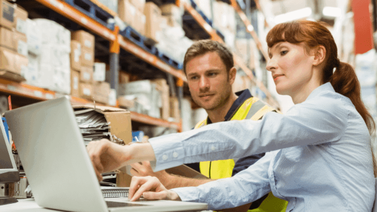 Two workers in a warehouse, one in a safety vest, sit at a table with a laptop. Shelves filled with boxes line the background as they review documents and collaborate on the computer.