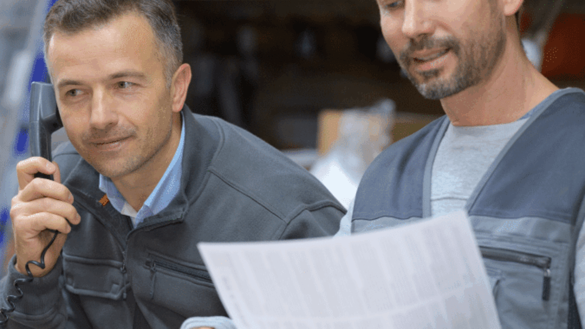 Two men in work uniforms are in a warehouse. One is talking on the phone while the other reviews a document, both appearing focused and engaged in their tasks. Boxes and shelves are visible in the background.