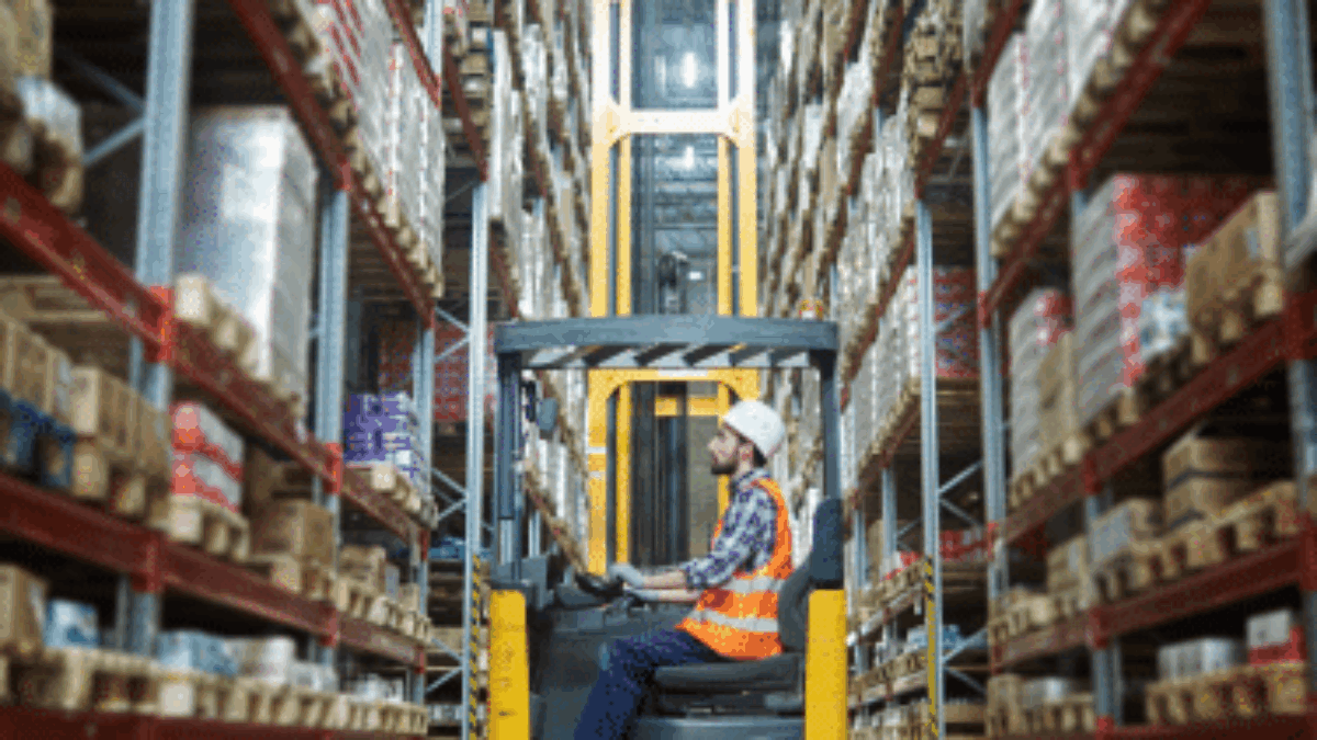 A worker in a safety vest and helmet operates a forklift between tall shelves stacked with boxes in a large warehouse.