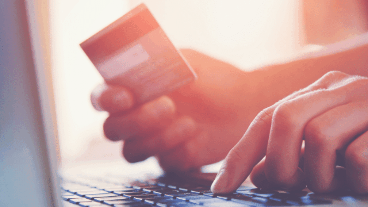 A person holds a credit card in one hand while typing on a laptop with the other, suggesting online shopping or electronic payment. The image is brightly lit with a soft background.
