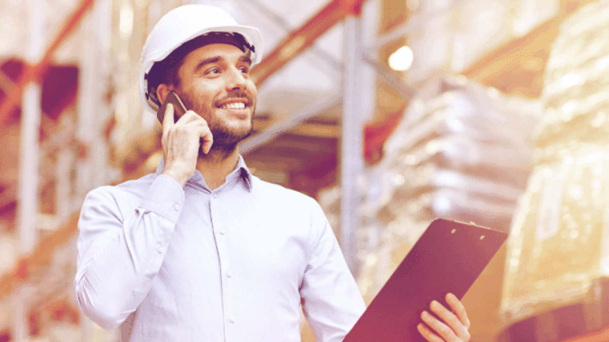 A man wearing a white safety helmet stands in a warehouse, smiling while talking on a phone and holding a clipboard. Shelves stocked with boxes and packages are visible in the background.