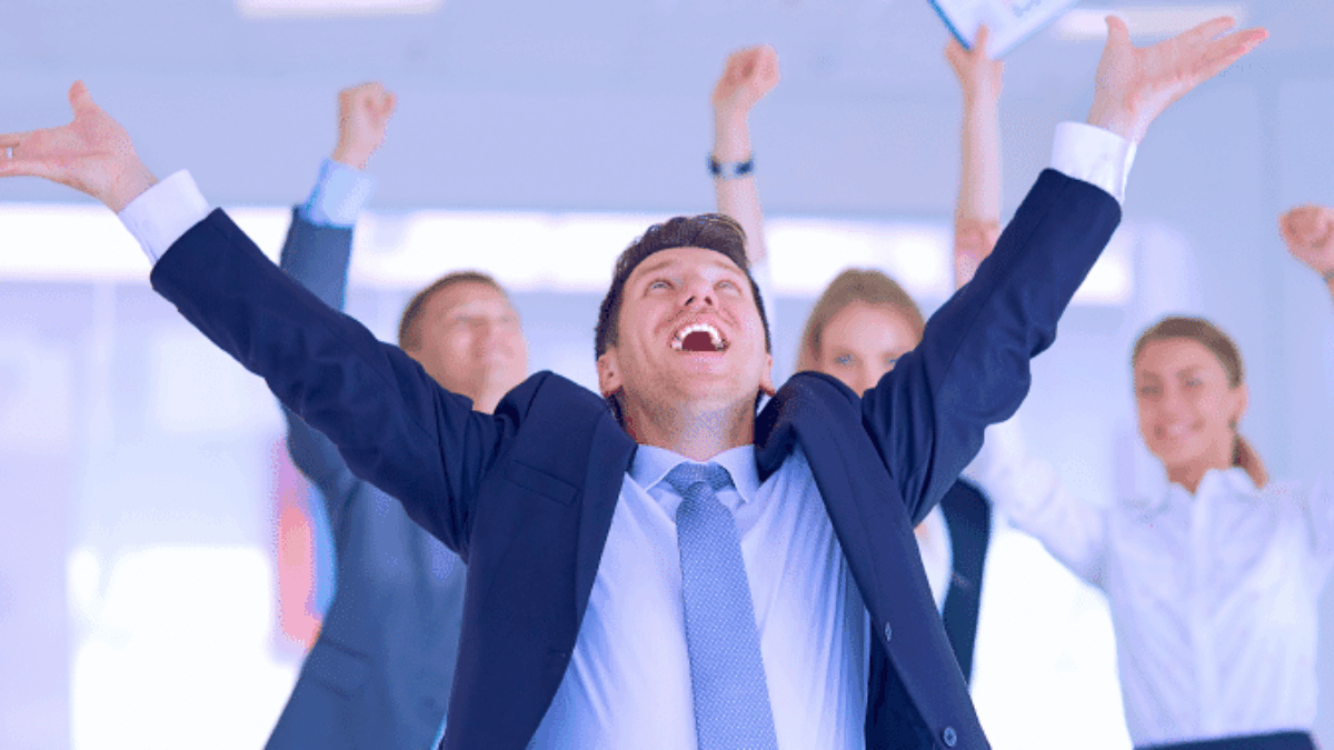 A group of businesspeople in formal attire celebrate enthusiastically in an office. One man stands at the front with arms raised and smiling broadly, while others cheer and raise their arms in the background.