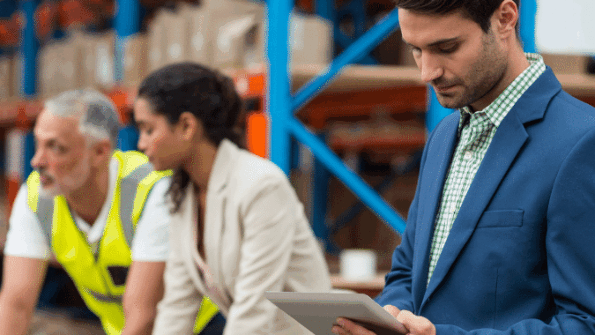 A man in a blue suit uses a tablet in a warehouse, while two colleagues, one in a safety vest and the other in business attire, review documents on a table in the background. Shelves with boxes are visible behind them.